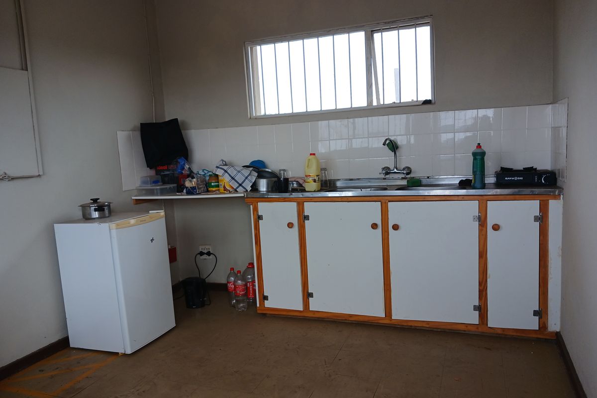 A basic industrial kitchenette featuring simple white cabinetry, a stainless steel sink, tiled splashback, and a window with security bars.