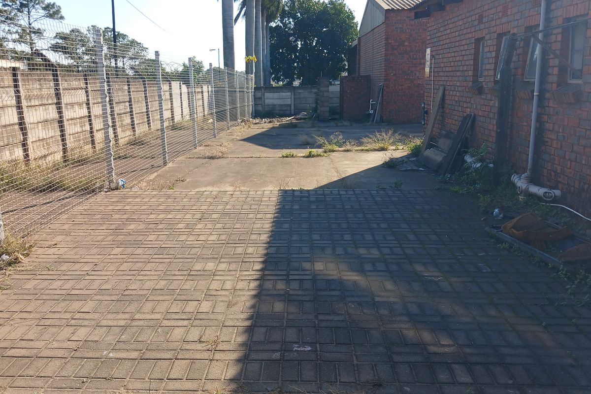 An exterior rear industrial yard area featuring paved surfaces, perimeter security fencing, and brick building facade with visible maintenance requirements.