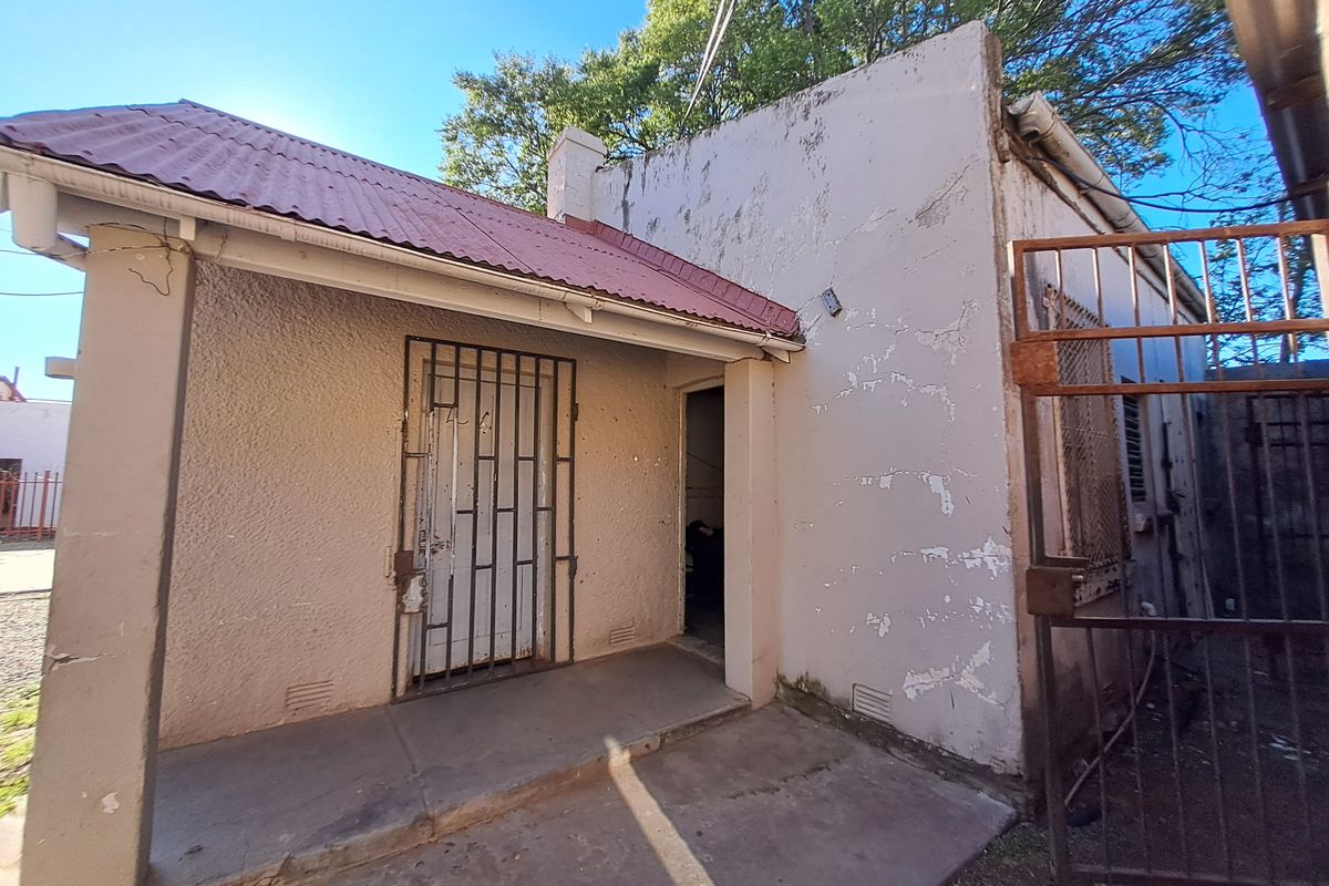 Rear exterior view of the property featuring a covered patio area, security gate, and weathered masonry walls requiring maintenance.