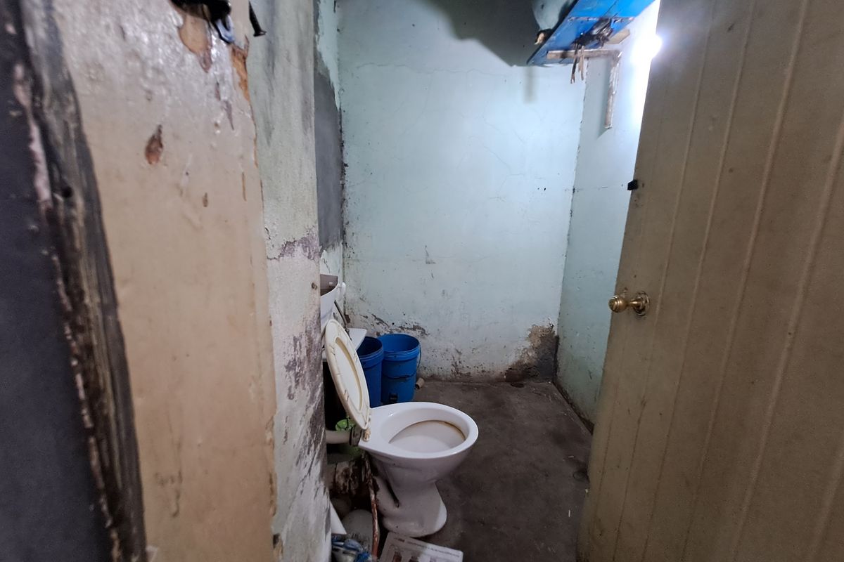A small, dilapidated bathroom space requiring significant renovation, featuring a basic toilet and showing extensive wear to wall finishes and door frames.