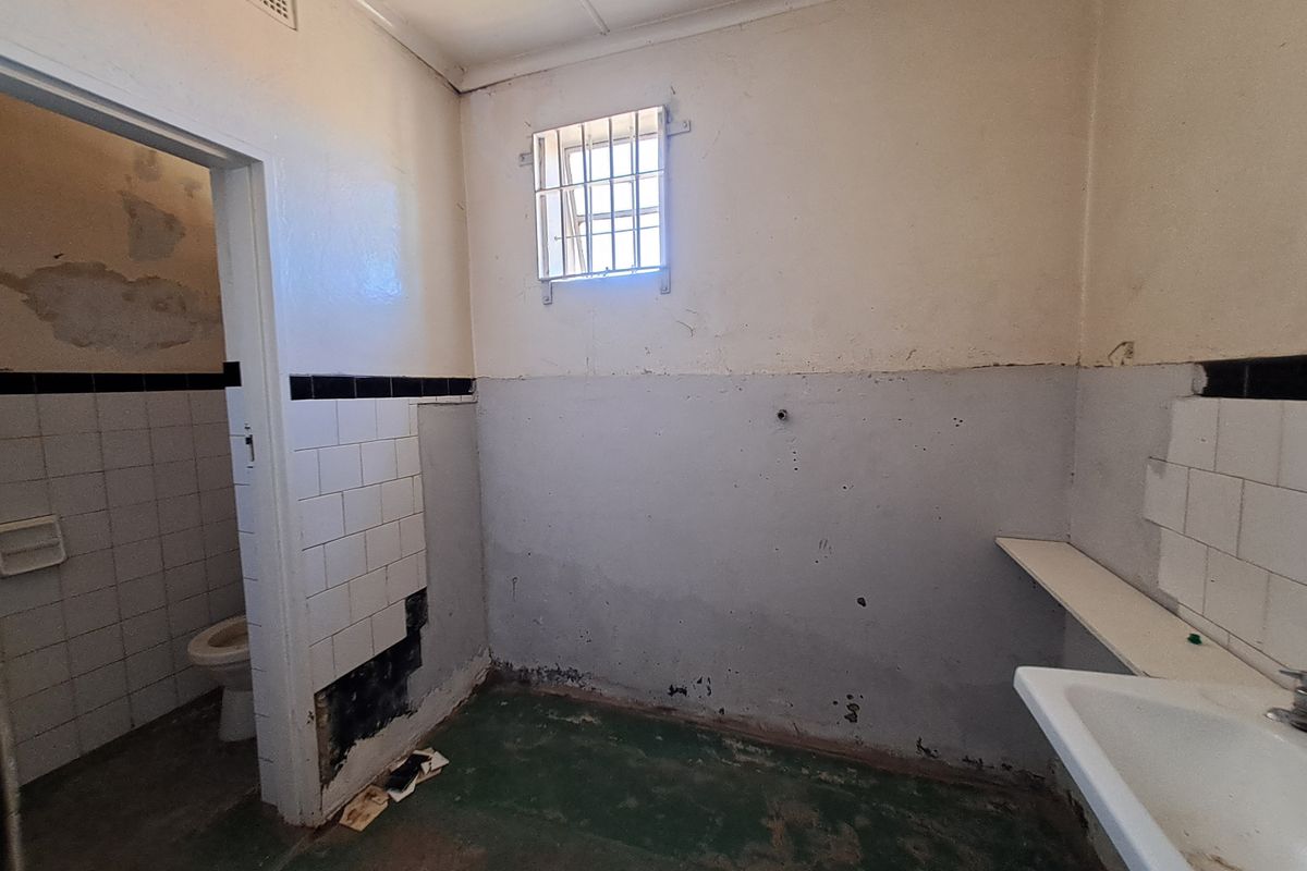 A vacant bathroom space requiring renovation, featuring a wall-mounted basin, toilet, and security-barred window with visible wear to wall finishes and flooring.