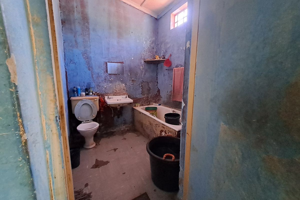A dilapidated bathroom requiring significant renovation, featuring a toilet, wall-mounted basin, and built-in bathtub with extensive wall and floor finish deterioration.