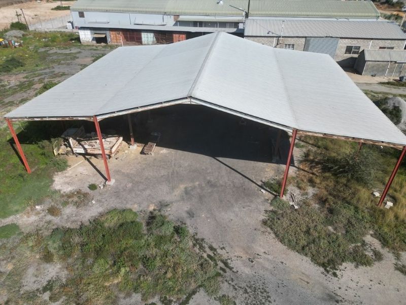 Aerial view of an open-sided industrial storage structure featuring a large corrugated metal roof supported by steel columns on an unpaved lot.