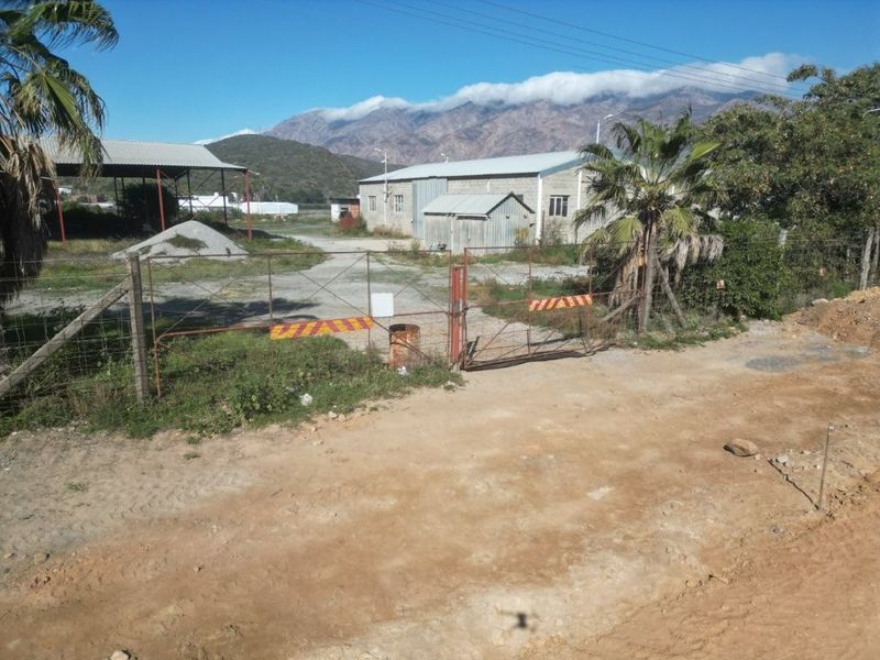 Aerial view of a commercial property featuring a gated entrance, unpaved driveway, and industrial-style buildings with corrugated metal roofing against a mountain backdrop.
