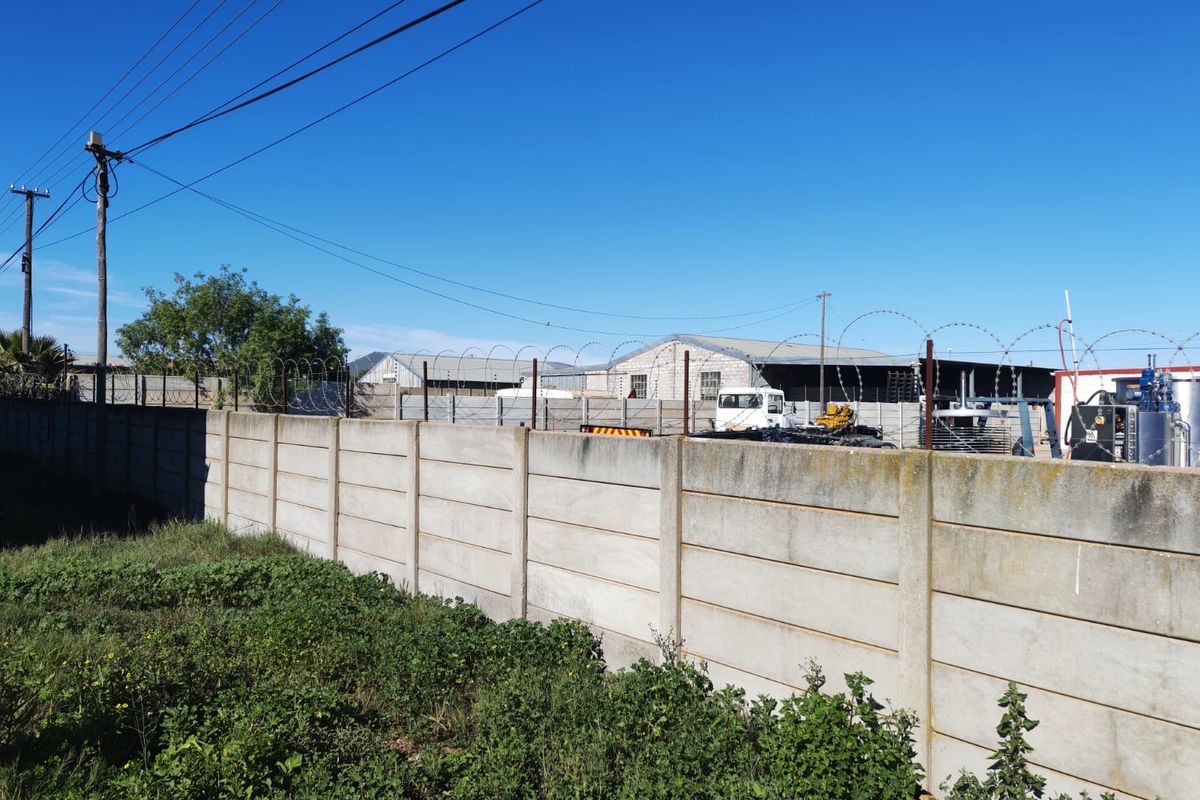 View of the industrial property boundary featuring a precast concrete wall with razor wire security fencing and adjacent warehouse structures.