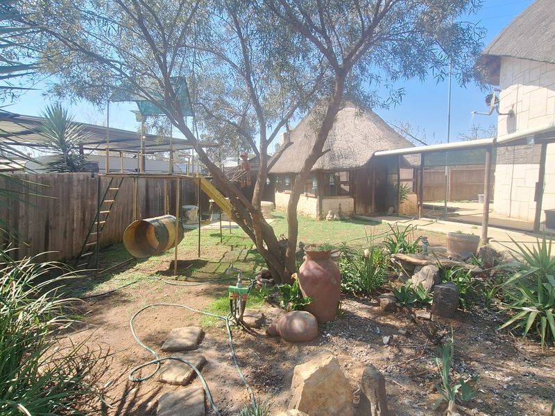 A spacious garden area featuring a thatched roof structure, mature trees, stone landscaping elements, and wooden perimeter fencing.