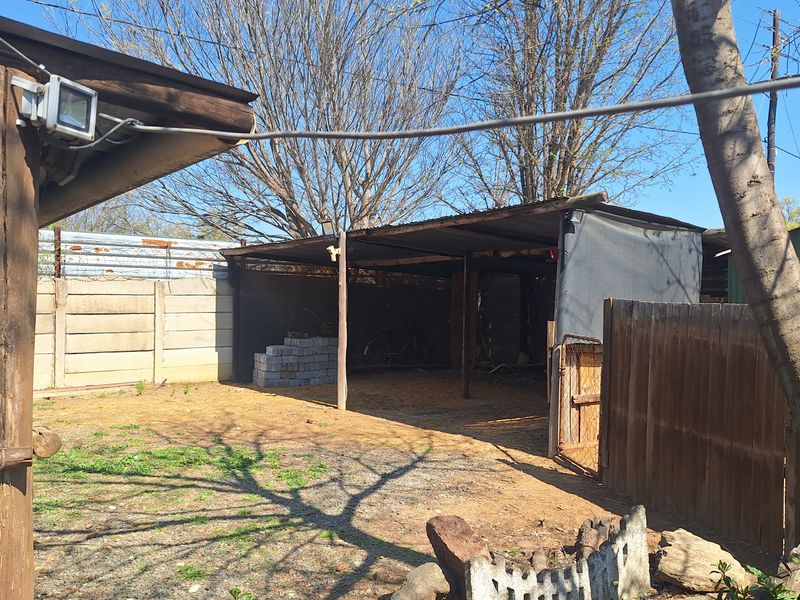 Rear outdoor area featuring a basic corrugated metal shelter, precast concrete boundary wall, and weathered wooden fencing on a gravel surface.
