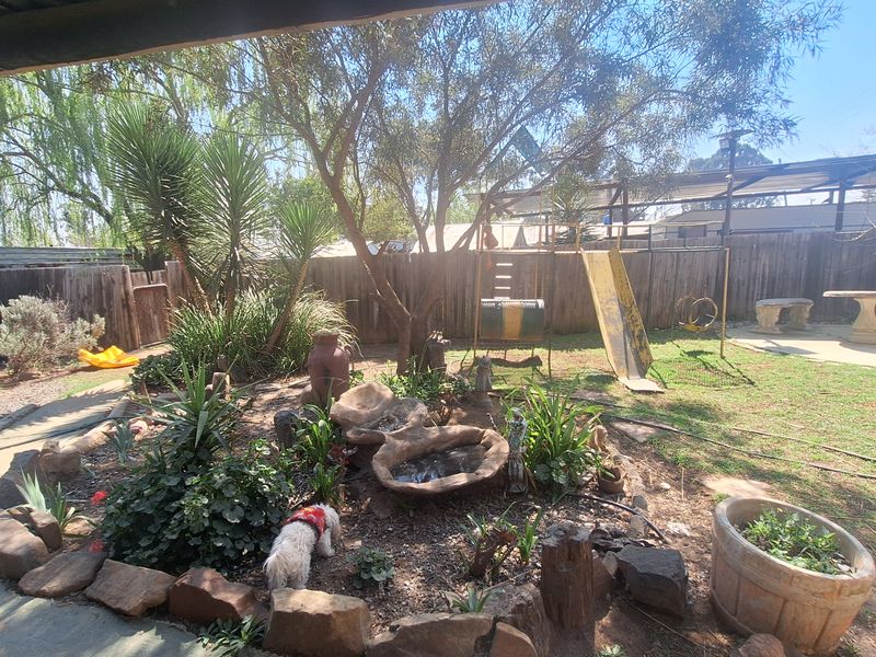 A backyard garden area featuring stone-edged landscaping, mature trees, and a covered patio space with timber fencing.