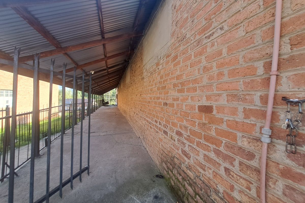 A covered exterior walkway featuring exposed brick walls, concrete flooring, and metal security fencing along the side of the commercial building.