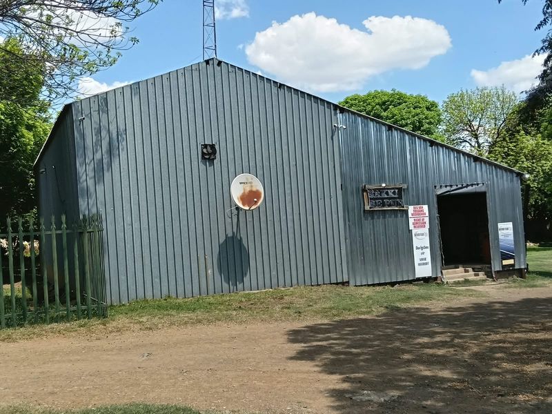 Exterior view of a commercial corrugated metal structure featuring a pitched roof, ventilation fan, and perimeter security fencing.