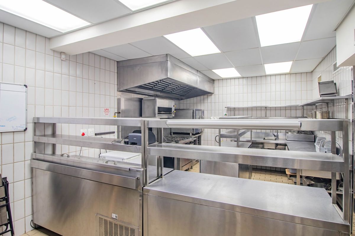 Commercial-grade kitchen featuring extensive stainless steel work surfaces, industrial extractor hood, and tiled wall finishes, designed for high-volume food preparation.