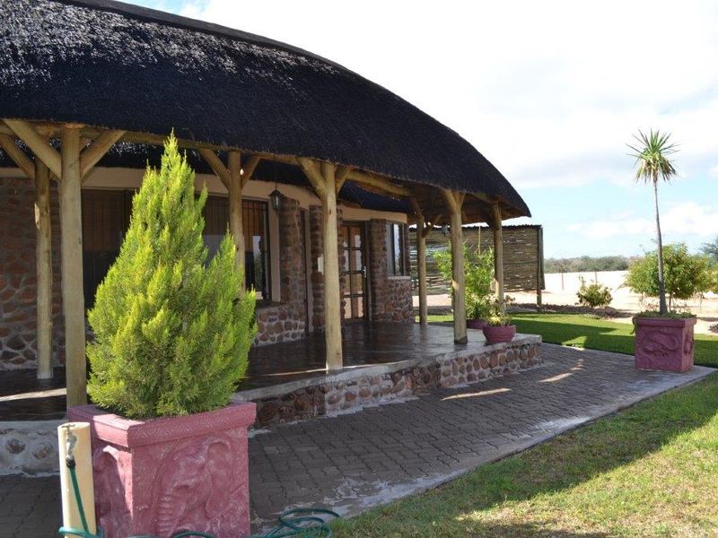 A well-maintained guest house exterior featuring a traditional thatch roof, timber support posts, and stone cladding with a spacious paved patio area.