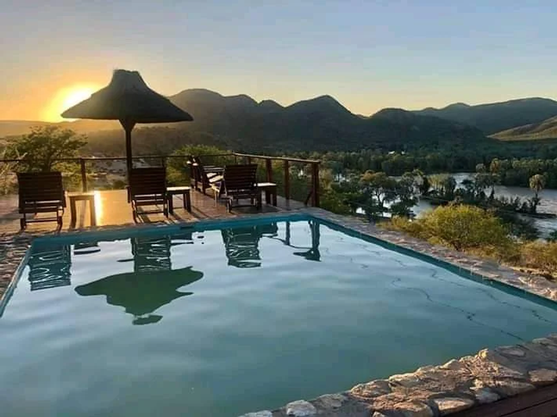An outdoor swimming pool area featuring stone coping and timber decking, offering panoramic views of the river and surrounding mountains at sunset.