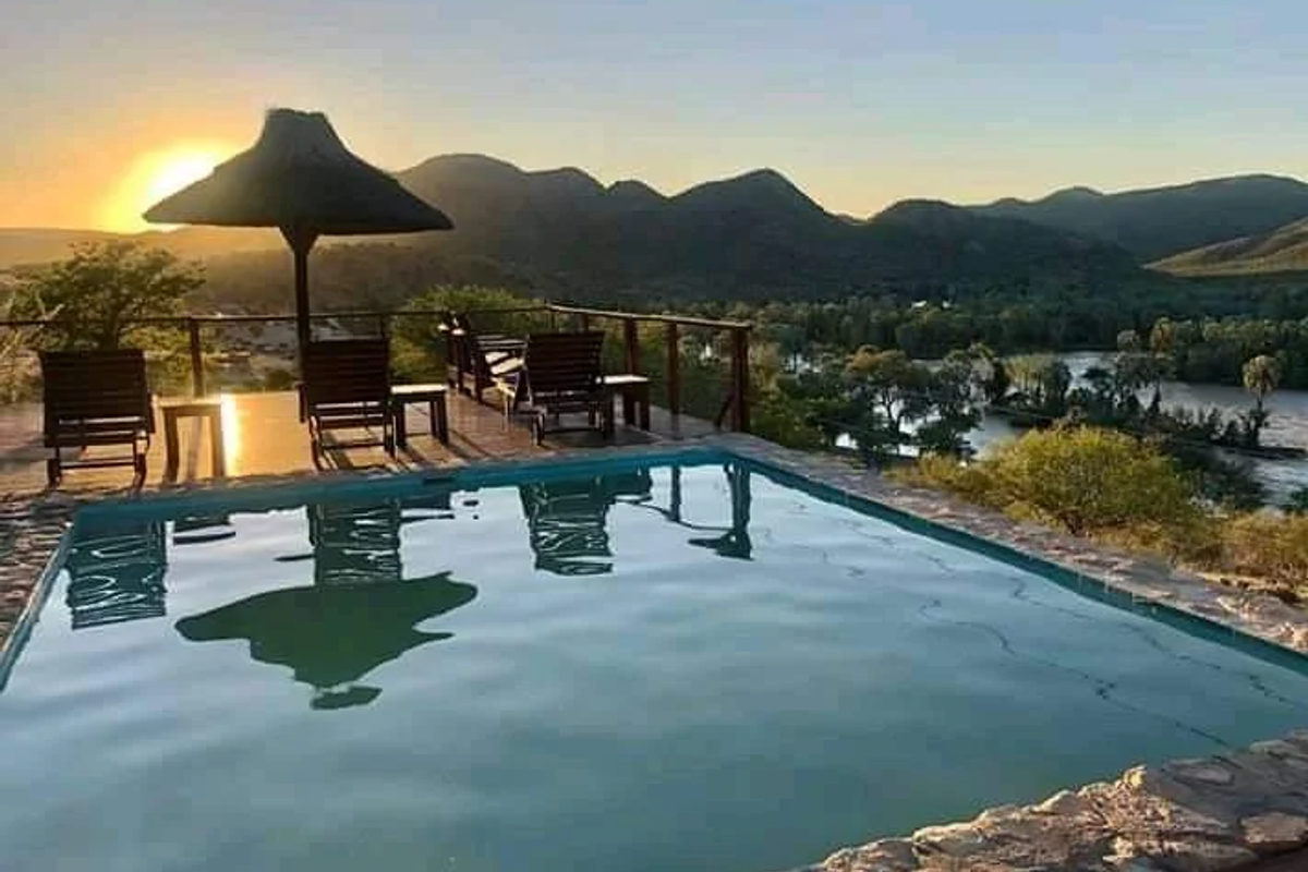 An outdoor swimming pool area featuring stone coping and timber decking, offering panoramic views of the river and surrounding mountains at sunset.