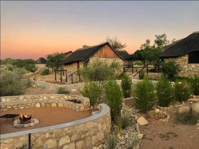A scenic outdoor area featuring stone-walled cottages with thatched roofs, a built-in fire pit, and landscaped garden paths at sunset.