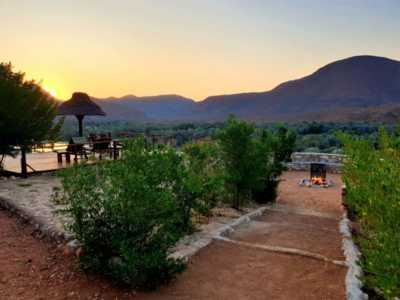Outdoor entertainment area featuring a wooden deck, stone fire pit, and thatched umbrella, offering panoramic mountain views at sunset.