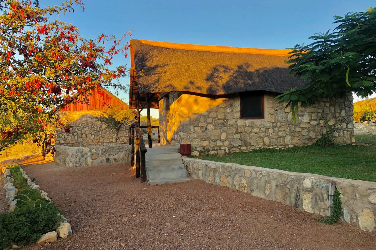 A rustic stone-clad building with a traditional thatched roof, featuring a concrete staircase and landscaped retaining walls in a natural setting.