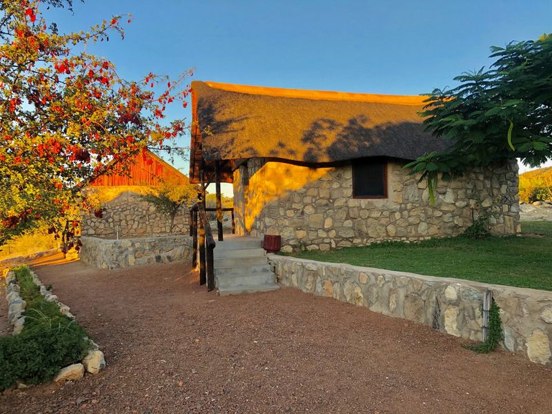 A rustic stone-clad building with a traditional thatched roof, featuring a concrete staircase and landscaped retaining walls in a natural setting.