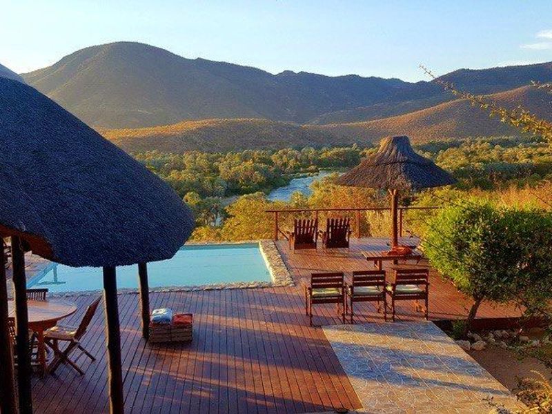 An outdoor pool area featuring timber decking, a thatched roof structure, and a swimming pool overlooking a scenic river and mountain landscape.