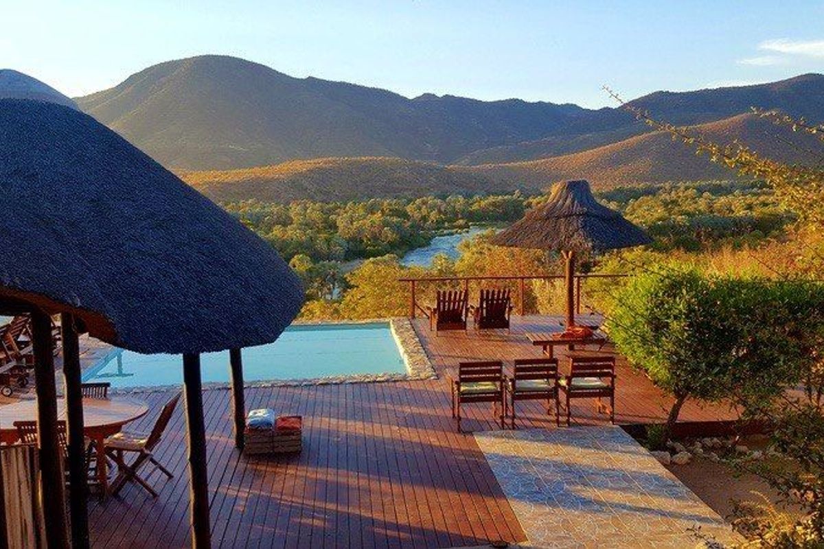 An outdoor pool area featuring timber decking, a thatched roof structure, and a swimming pool overlooking a scenic river and mountain landscape.