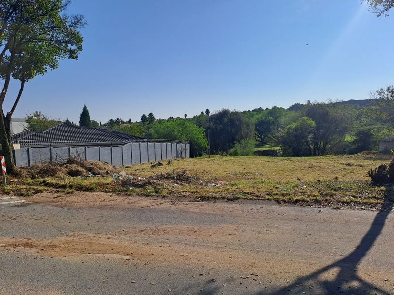 A vacant residential plot featuring a boundary wall, accessible from a paved road, with surrounding mature trees and greenery.