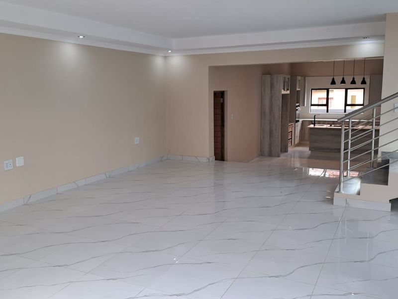 Spacious open-plan living area featuring modern white marble-look floor tiles, recessed lighting, and a contemporary staircase with stainless steel railings leading to the kitchen.