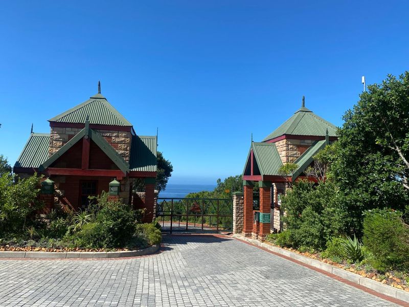 A well-maintained gated entrance featuring stone gatehouses, metal roofing, and a paved driveway leading toward a scenic ocean view.