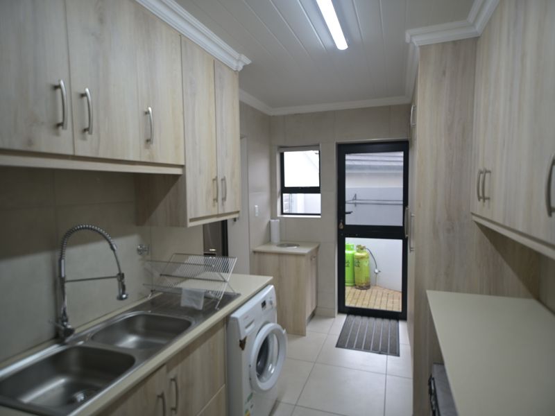 A functional laundry room featuring light wood-look cabinetry, double stainless steel sink, tiled splashbacks, and direct access to an outdoor area.