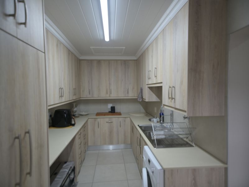 A functional scullery featuring light wood-finish cabinetry, stone countertops, a stainless steel sink, and tiled flooring with efficient overhead lighting.