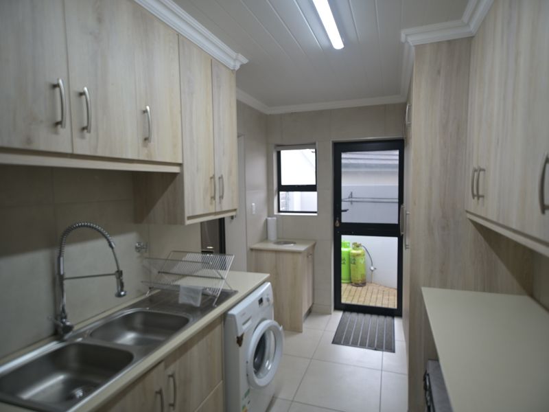 A functional laundry room featuring light wood-finish cabinetry, a double stainless steel sink, tiled flooring, and direct access to an outdoor area.