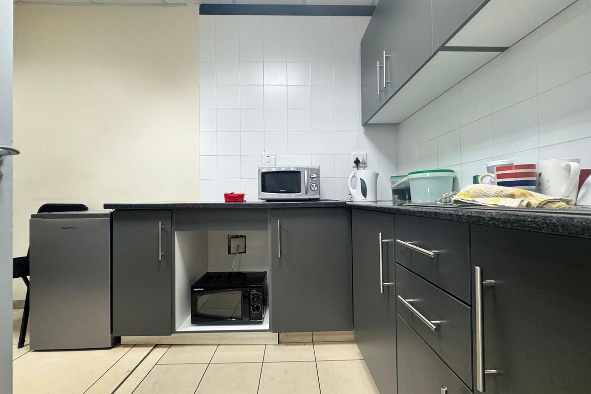A functional office kitchenette featuring dark grey cabinetry, granite-look countertops, white tiled splashbacks, and a drop ceiling.