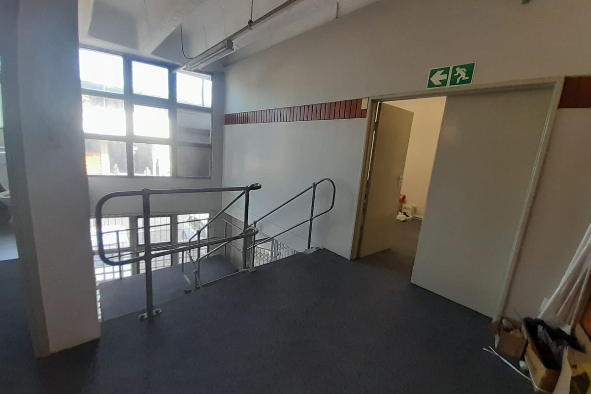 Industrial hallway featuring a staircase with metal handrails, large window panels, and emergency exit signage, providing access to adjacent office or utility spaces.
