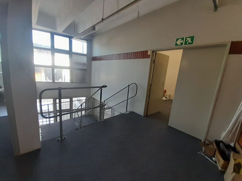 Industrial hallway featuring a staircase with metal handrails, large window panels, and emergency exit signage, providing access to adjacent office or utility spaces.