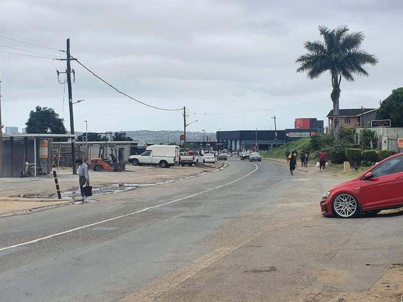 A street-level view of a commercial area featuring asphalt road infrastructure, utility poles, and perimeter fencing along the roadside.