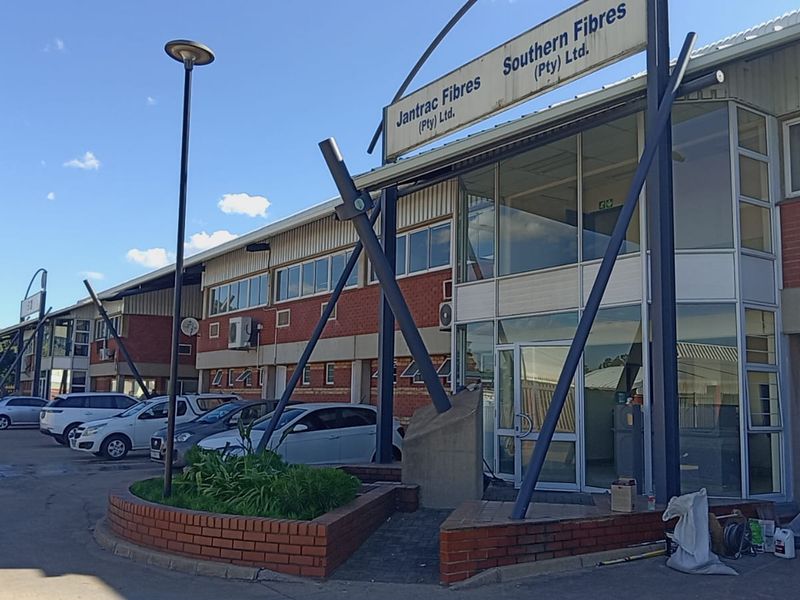 Front elevation of an industrial facility featuring brick construction, large glass windows, and prominent signage, with an adjacent parking area.