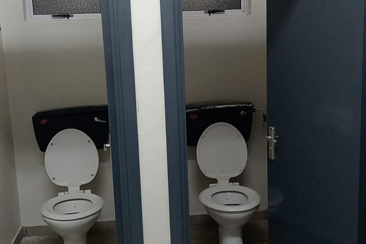 A functional industrial bathroom facility featuring two separate toilet cubicles with tiled flooring and security-barred windows.