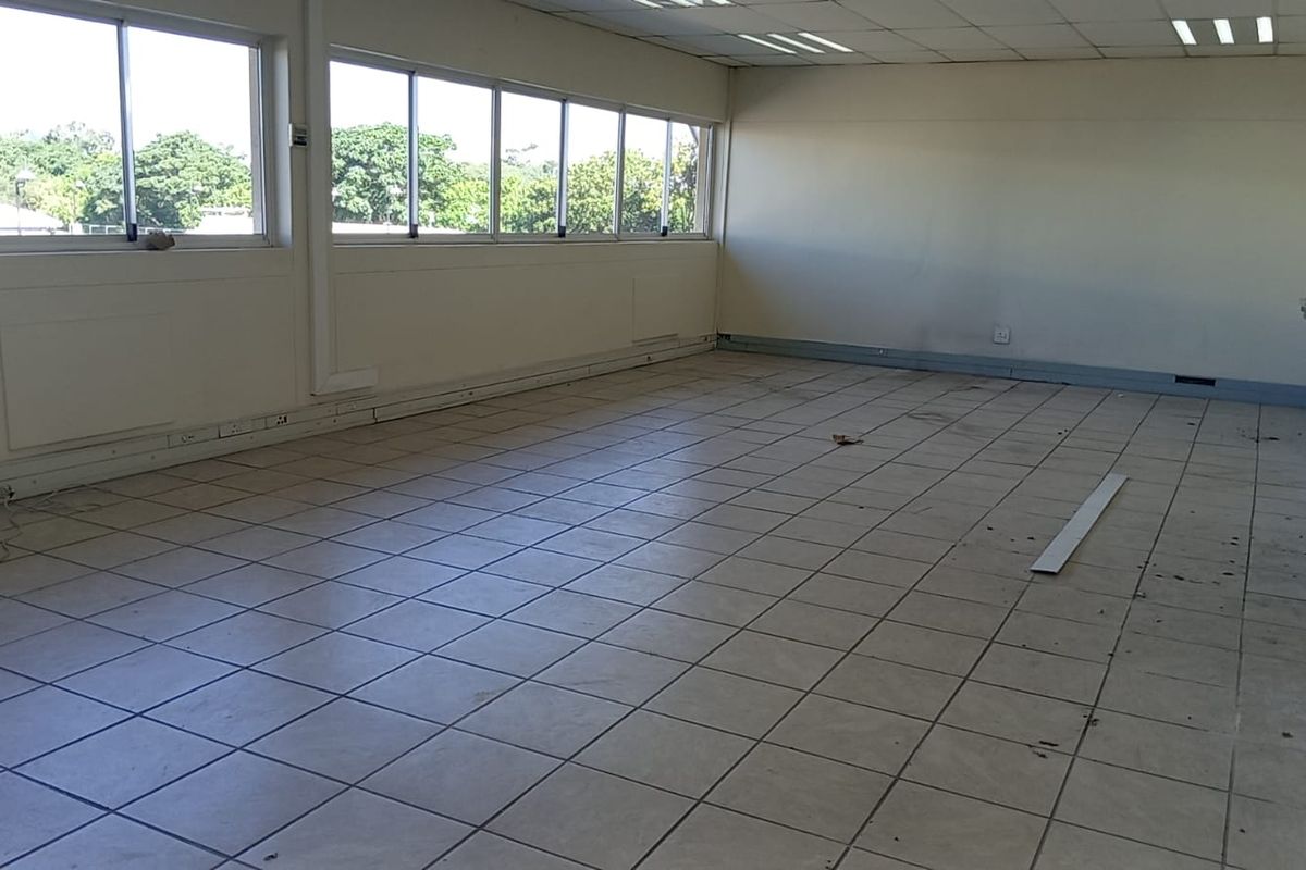 An empty, open-plan industrial office space featuring tiled flooring, large windows for natural light, a suspended ceiling, and a wall-mounted sink.