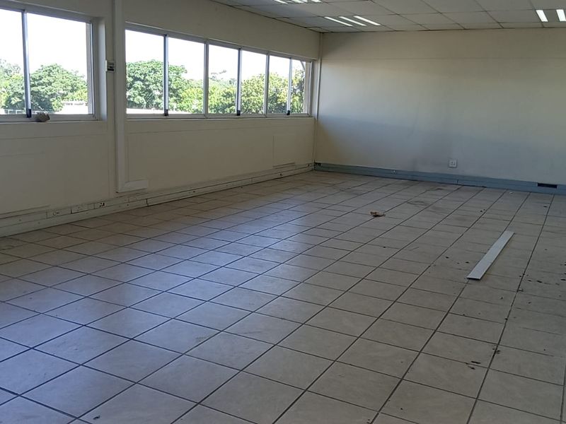 An empty, open-plan industrial office space featuring tiled flooring, large windows for natural light, a suspended ceiling, and a wall-mounted sink.