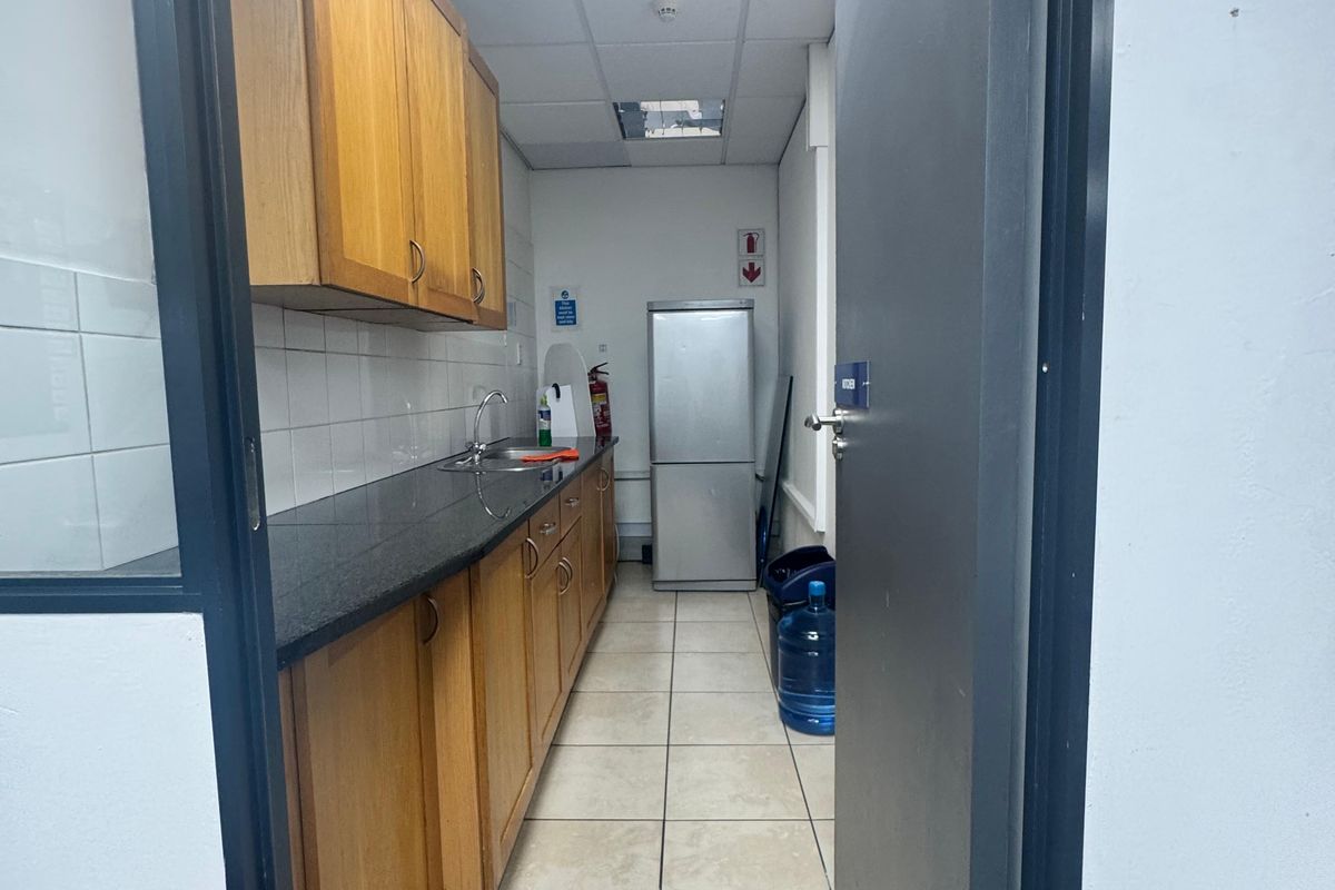 A functional office kitchenette featuring wooden cabinetry, a dark countertop, a stainless steel sink, and tiled flooring.