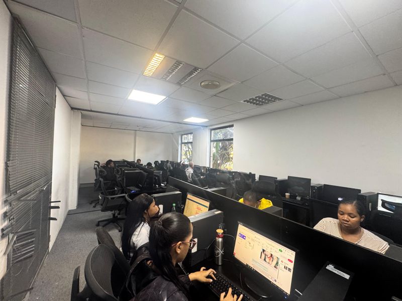 A functional office space featuring rows of built-in workstations, suspended ceiling tiles, and large windows providing natural light.