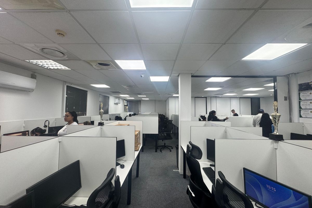 Open-plan office space featuring multiple partitioned workstations, suspended ceiling grid with recessed lighting, and wall-mounted air conditioning unit.