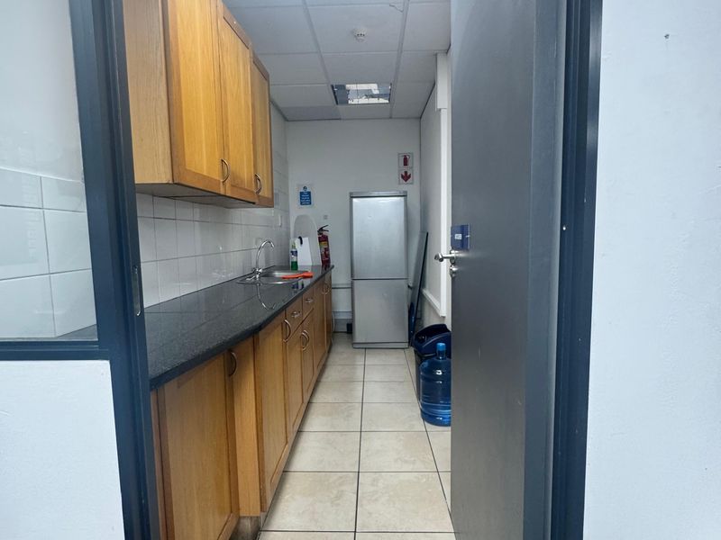 A functional office kitchenette featuring wooden cabinetry, a dark countertop, a stainless steel sink, and tiled flooring.