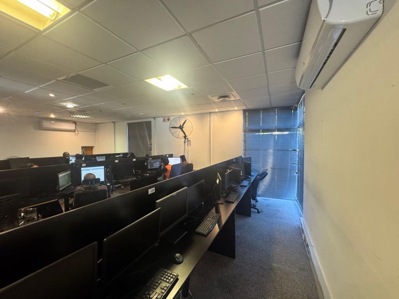 Open-plan office space featuring suspended ceiling tiles, wall-mounted air conditioning units, integrated desk workstations, and window blinds for light control.
