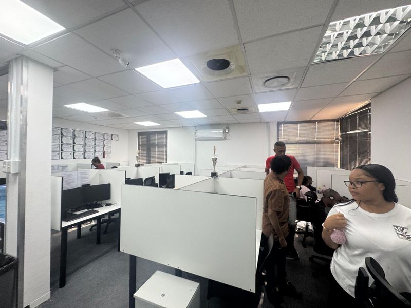 Open-plan office space featuring built-in workstation cubicles, suspended acoustic ceiling tiles, integrated air conditioning unit, and window blinds.