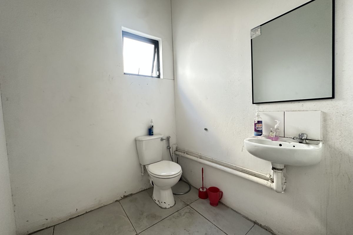 A functional industrial-style bathroom featuring a wall-mounted basin, toilet, and mirror with exposed plumbing and tiled flooring.