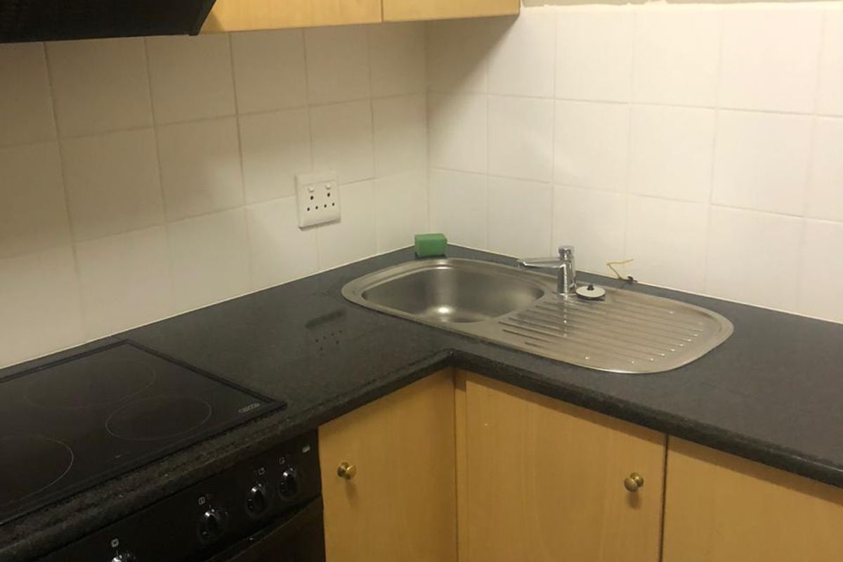 Compact office kitchenette featuring light wood cabinetry, dark countertops, a built-in electric stove, and a stainless steel sink with tiled splashback.