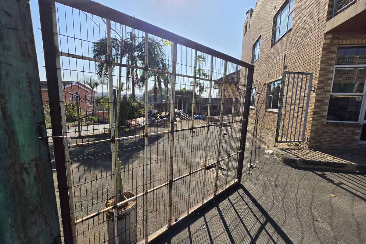 Rear exterior view of a commercial property featuring a paved yard, wire mesh fencing, and brick building facade.
