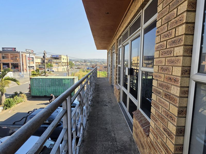 An exterior balcony featuring face brick walls, aluminium window frames, and a metal balustrade overlooking the street and surrounding commercial area.