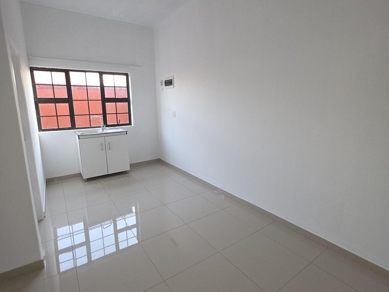A clean, empty kitchen area featuring tiled flooring, a single basin sink with base cabinetry, and large windows with security bars.