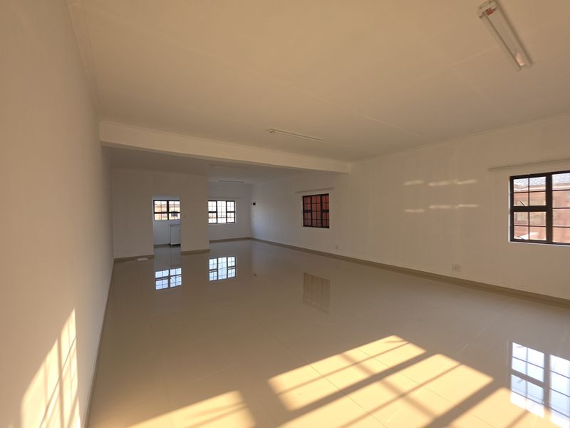 A spacious, empty open-plan living area featuring neutral tiled flooring, ample natural light from grid-pane windows, and clean white walls.